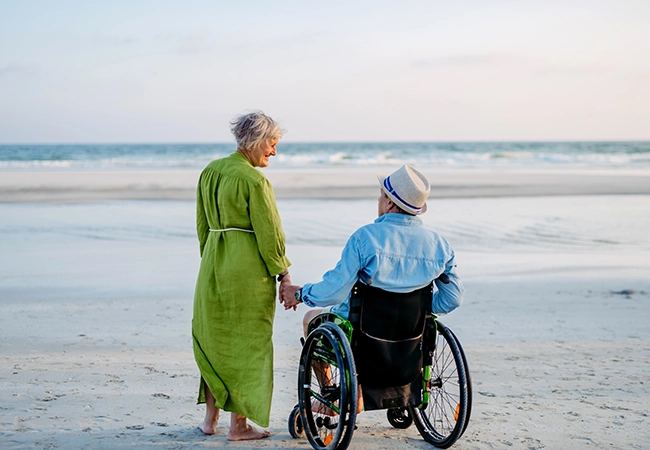 Image of woman standing next to a man sitting in a wheelchair at the beach Image of woman standing next to a man sitting in a wheelchair at the beach